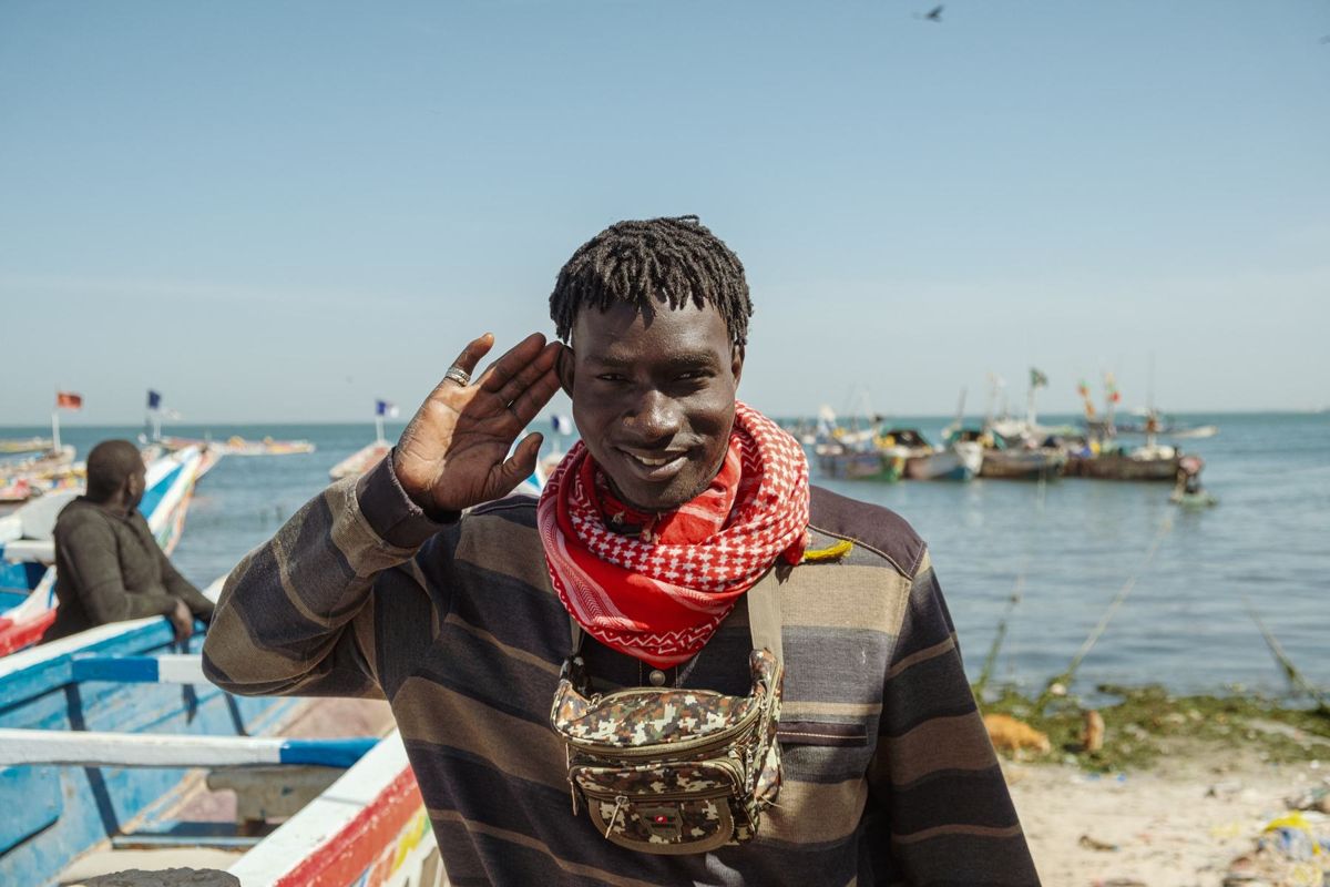 A smiling man in a red scarf raising his hand in greeting in front of fishing boats