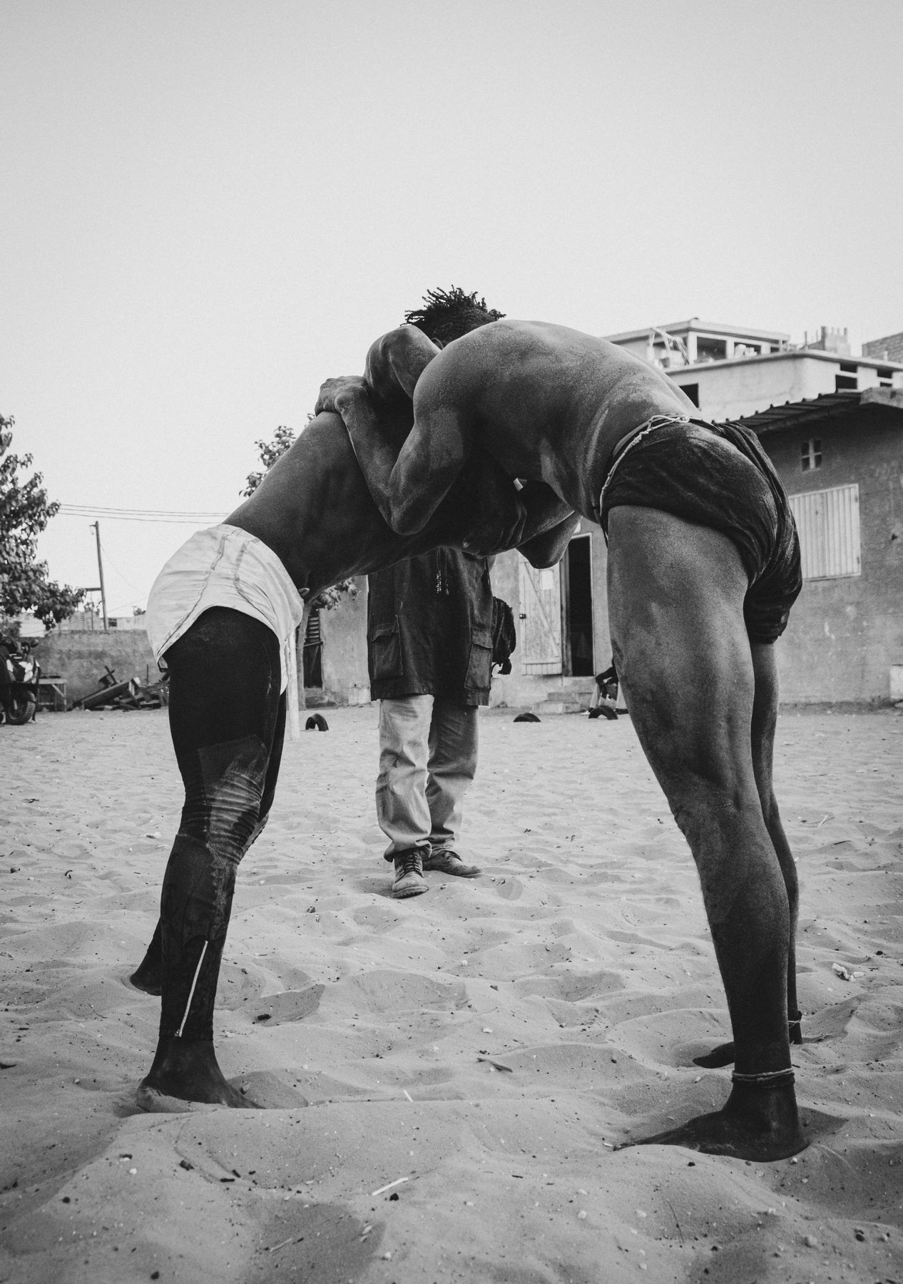 Two wrestlers locked head to shoulder in the sand while a man watches behind them