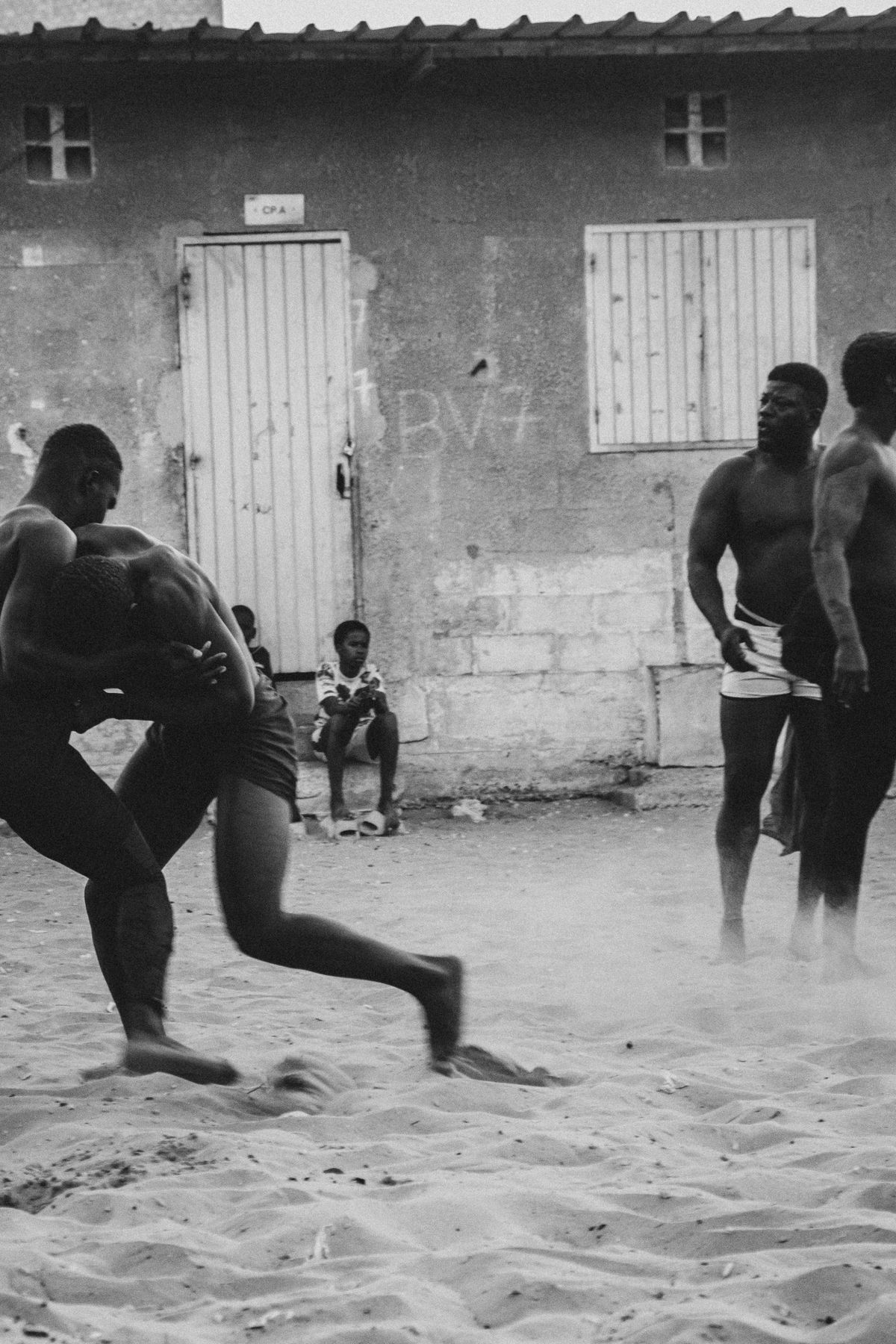 Two wrestlers in motion raising dust, a boy sitting by the door
