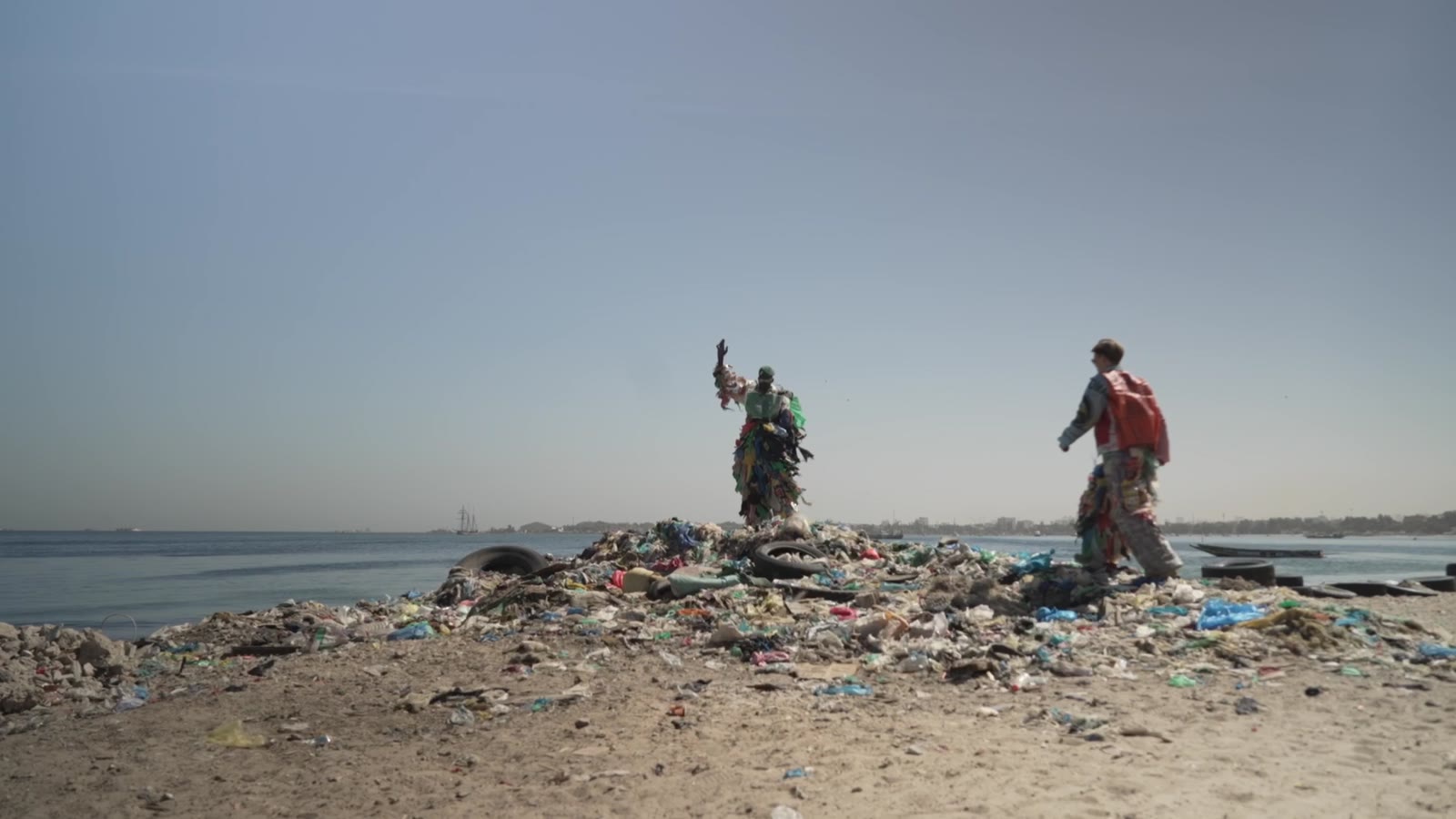 Modou Fall and Niklas Bruggeman in their plastic costumes standing on a heap of plastic waste by the sea