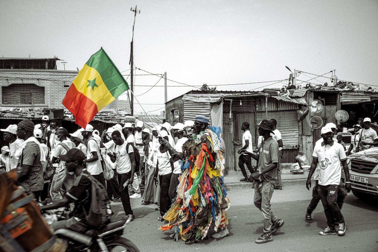 L’Homme Plastique in a plastic costume marches with a Senegalese flag through a crowd in white caps