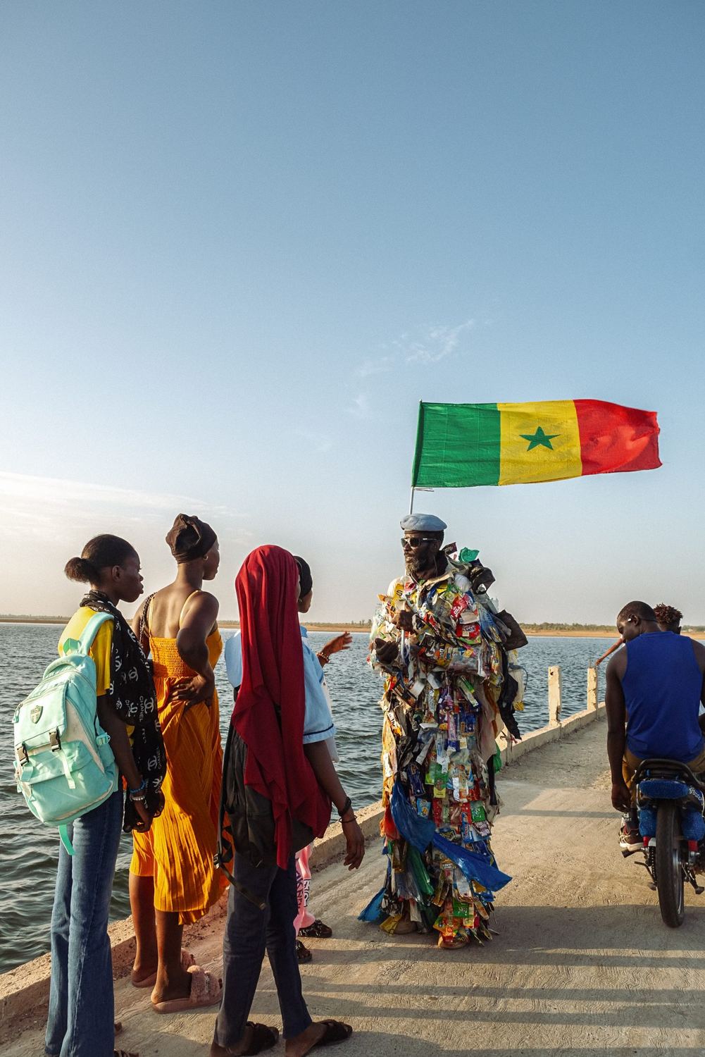 L’Homme Plastique talks with young people on a jetty, a Senegalese flag above him