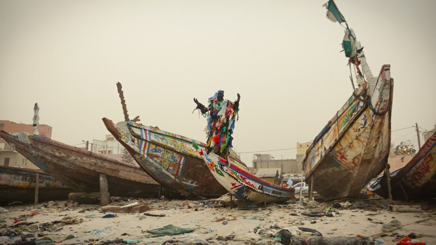 L’Homme Plastique stands with raised arms on a washed-up pirogue among rubbish on the beach