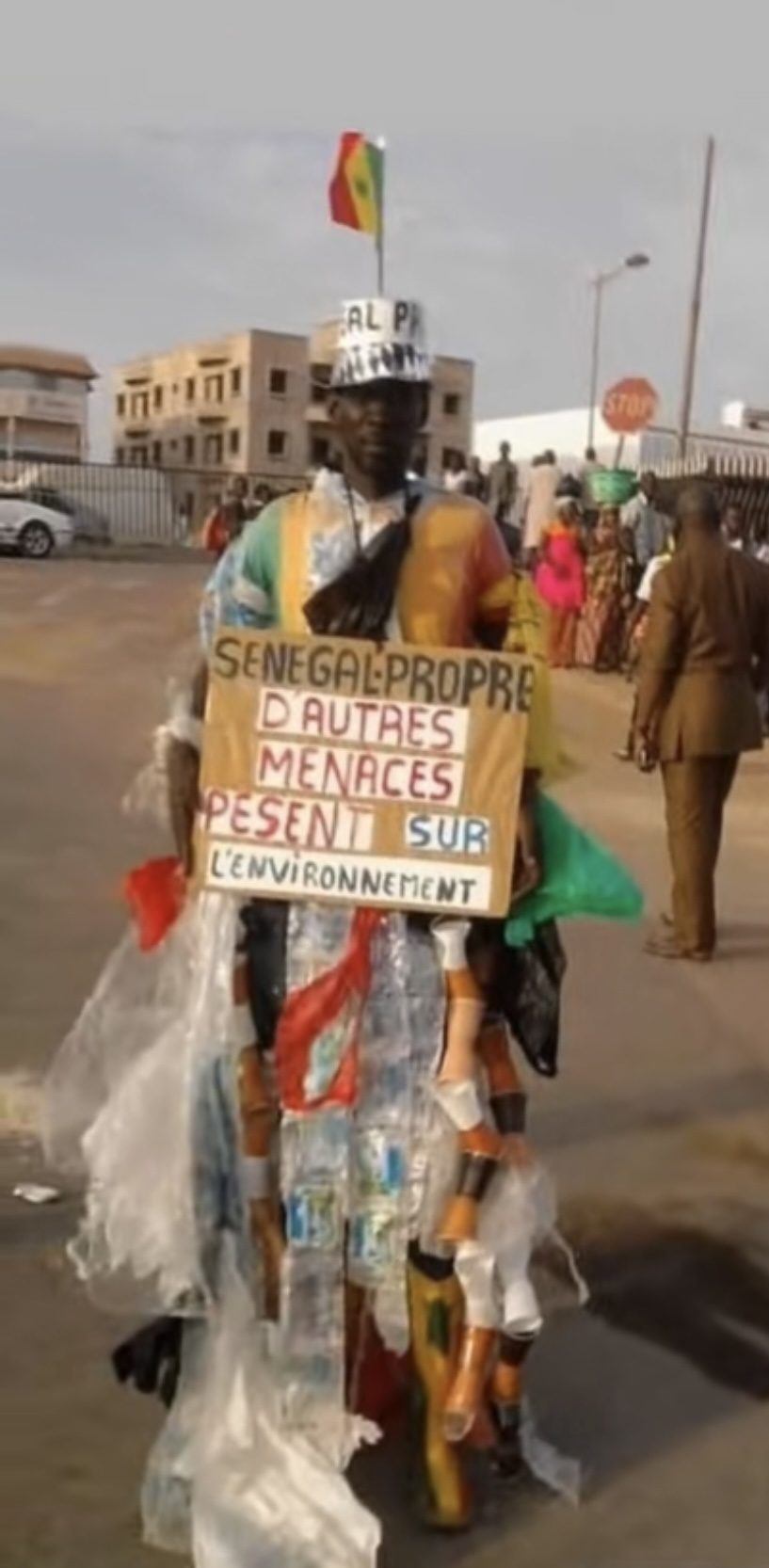 Modou Fall, L’Homme Plastique, in a costume of plastic bottles and bags, holding a cardboard sign that reads Sénégal Propre, d’autres menaces pèsent sur l’environnement